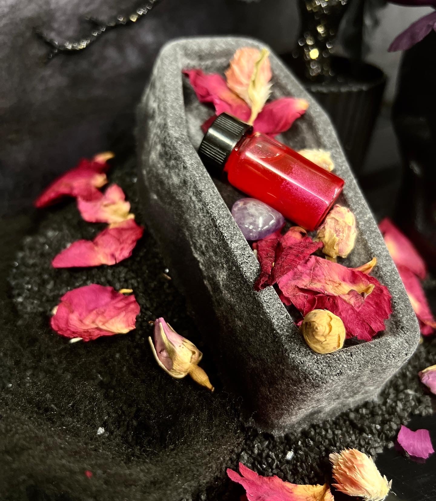 one cauffin shaped bath bomb laying down with rose petals and small vial of pink oil and purple amethyst stone 