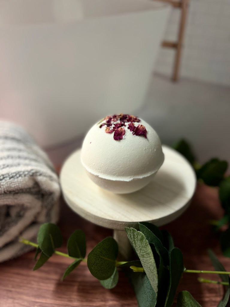 one singular white bath bomb with rose petals on top, sitting on a wooden pedestal in front of a fake bathroom backdrop with a gray and white towel rolled up next to it on the left.