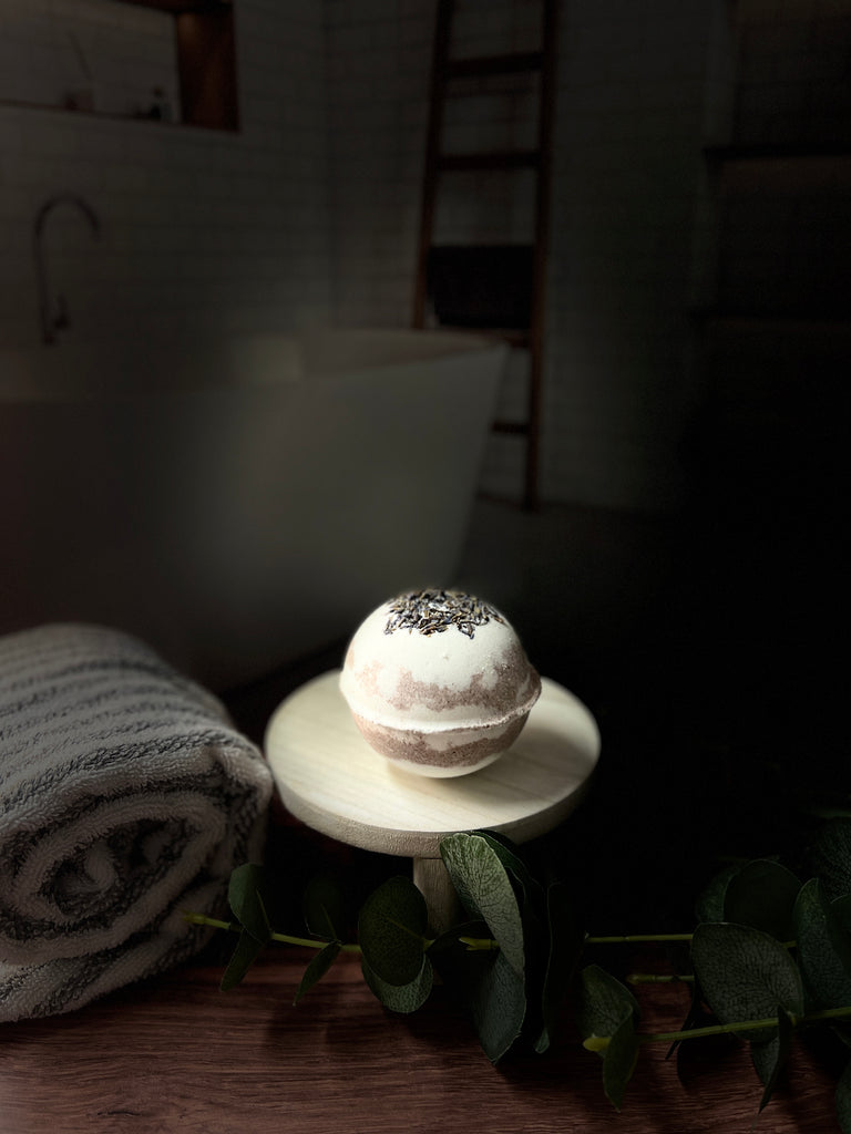 one singular purple and white bath bomb with lavender buds on top, sitting on a wooden pedestal in front of a fake bathroom backdrop with a gray and white towel rolled up next to it on the left.