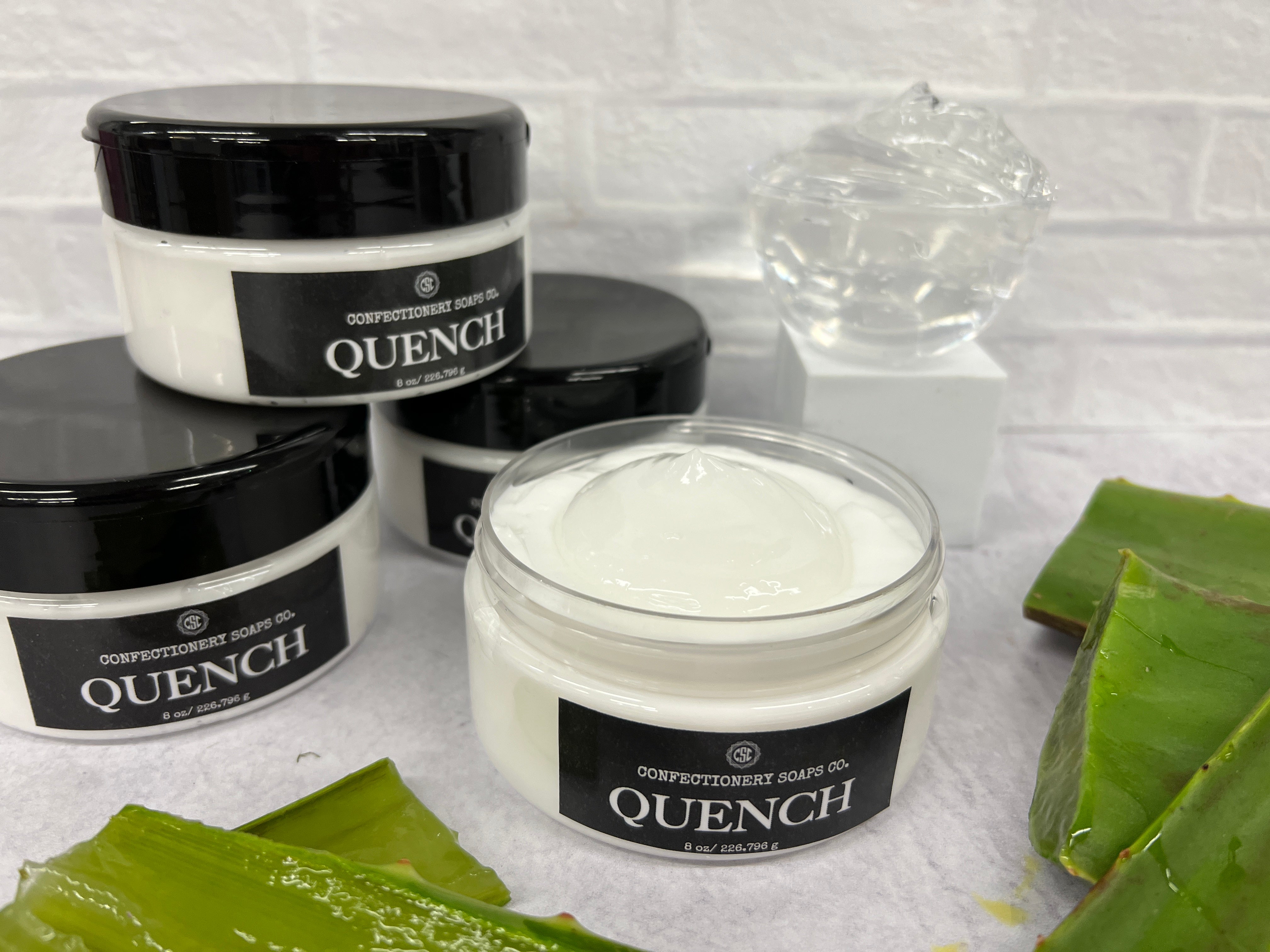 three quench jars stacked in a triangular formation for height, an open/exposed quench jar so you can see the aloe center, a bowl of aloe gel as deocration and aloe plant pieces surrounding the quench jars as decoration.